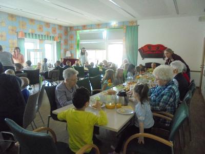 Senioren und Kinder sitzen gemeinsam im Veranstaltungsraum im Mehrgenerationenhaus sOfA am Tisch und genießen das Osterfrühstück.