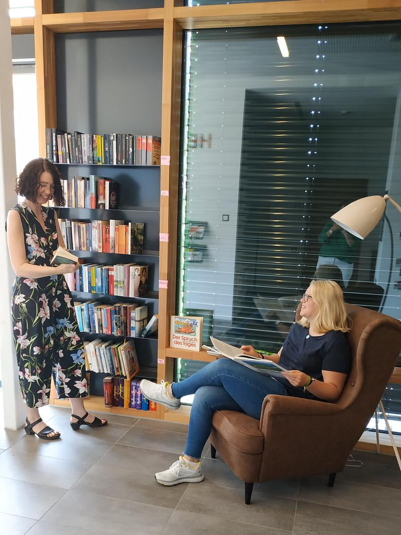 Das Bücherregal im Mehrgenerationenhaus sOfA. Es ist fast vollständig mit Büchern gefüllt. Eine Frau steht mit einem Roman in der Hand davor, eine andere sitzt auf einem Sessel daneben mit einem aufgeschlagenen Bildband auf dem Schoß.