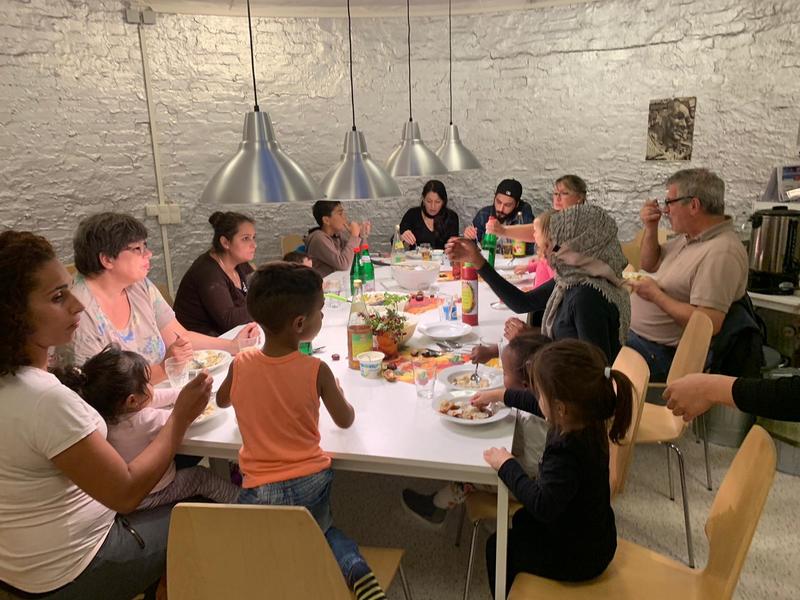 Frauen, Männer und Kinder verschiedener Nationen sitzen um einen Tisch und essen selbst gekochte Pelmeni.